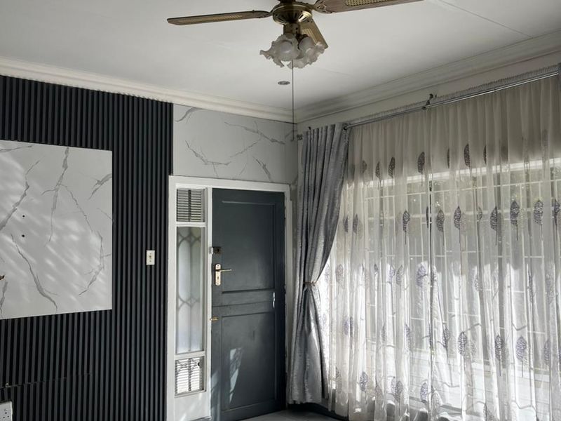 A bright living room featuring tiled flooring, decorative wall paneling, and a ceiling fan, with natural light streaming through large windows.