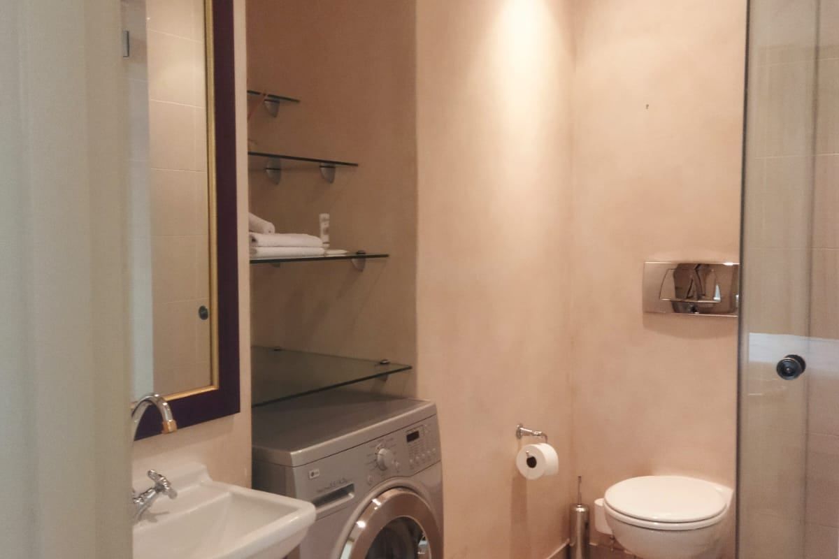 Compact bathroom featuring a wall-mounted toilet, glass shower enclosure, pedestal basin, and integrated laundry space with shelving.