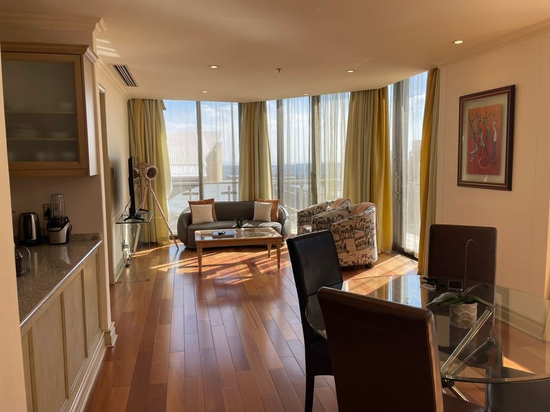 Spacious open-plan living and dining area featuring hardwood flooring, floor-to-ceiling windows with natural light, and integrated kitchen cabinetry.
