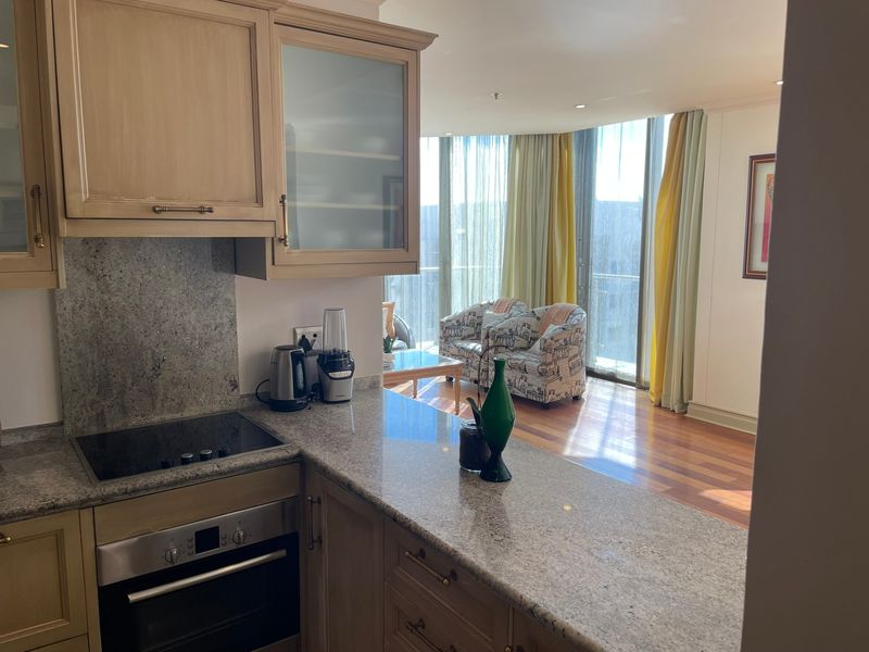 Open-plan kitchen featuring granite countertops, built-in oven, and hob, flowing into a bright living area with hardwood floors and floor-to-ceiling windows.