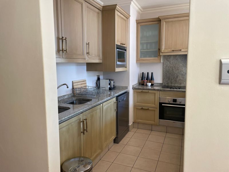 A functional kitchen featuring granite countertops, built-in oven, electric hob, integrated dishwasher, and ample cabinetry, finished with neutral tiled flooring.