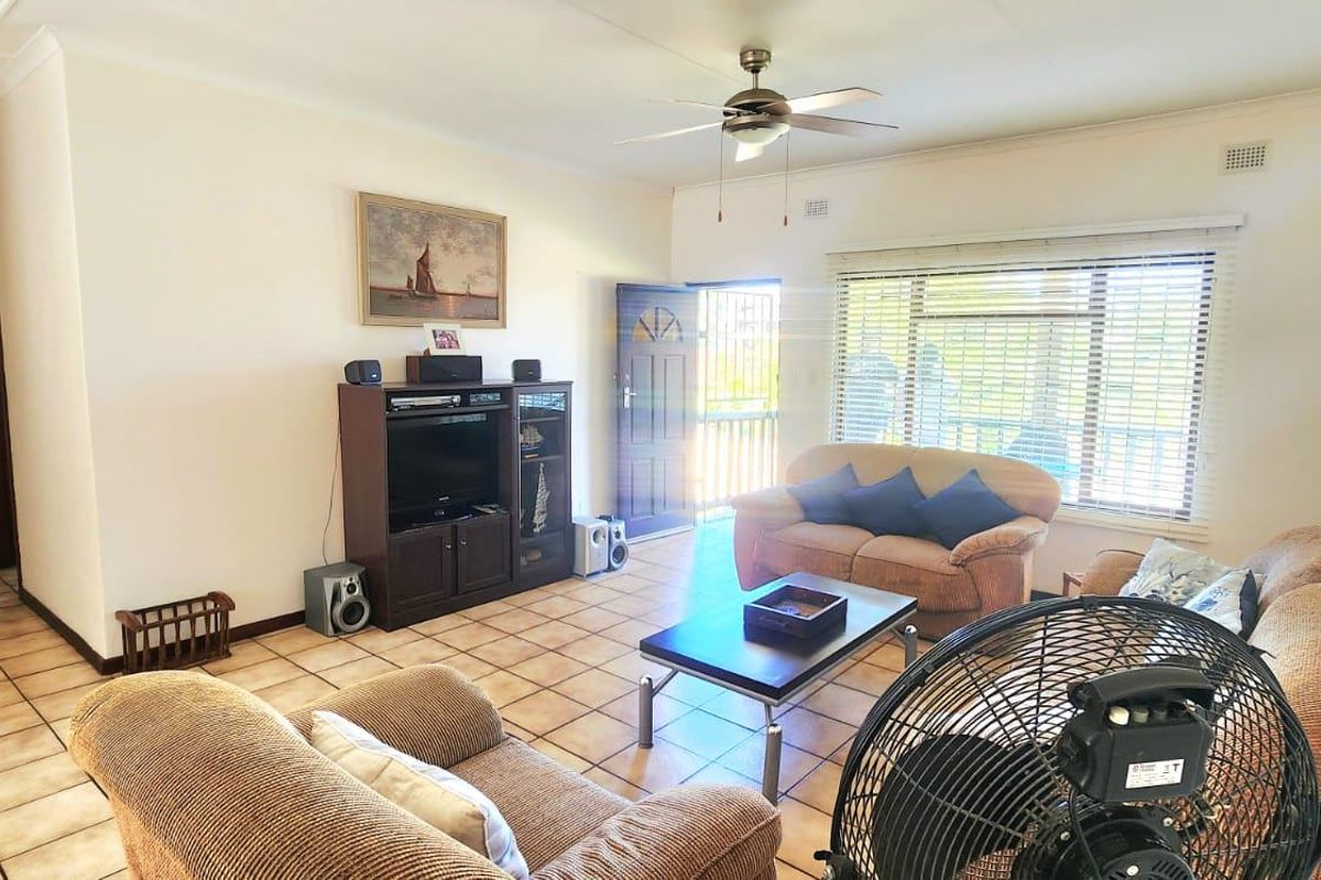A spacious living room featuring tiled flooring, a ceiling fan, and natural light through a large window with access to an exterior security gate.