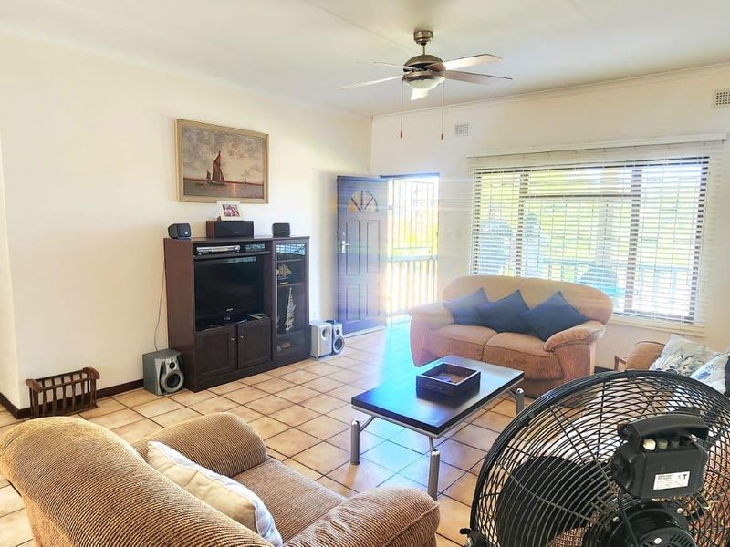 A spacious living room featuring tiled flooring, a ceiling fan, and natural light through a large window with access to an exterior security gate.