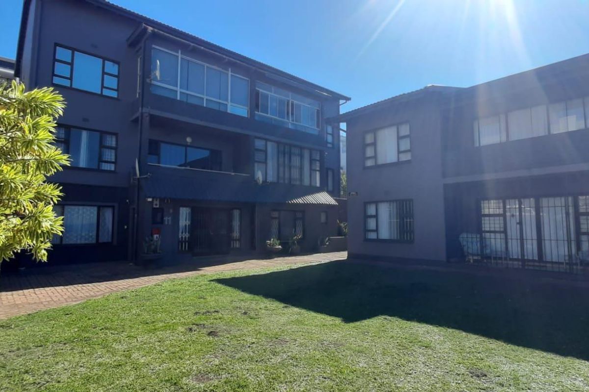 Rear view of the apartment complex featuring a shared lawn area, paved courtyard, and multi-story building facade with security-fitted windows and doors.