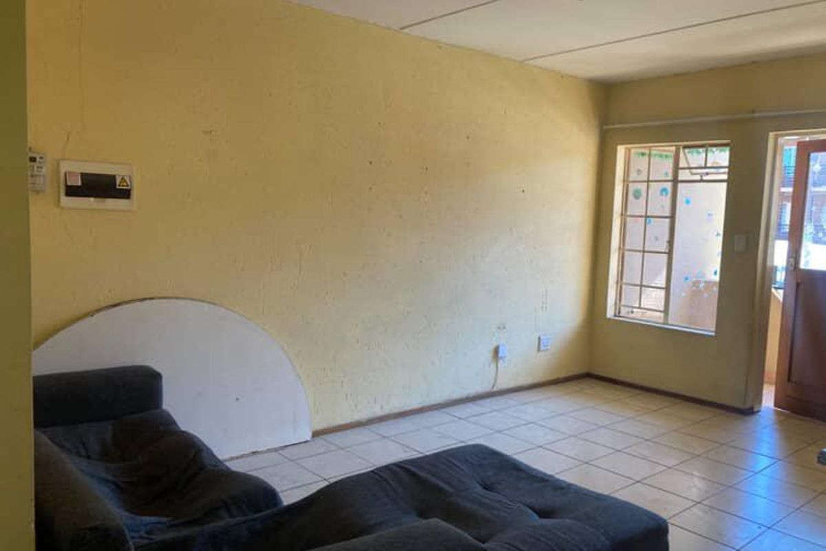 A living room area featuring tiled flooring, a security gate, and a wall-mounted electrical distribution board, requiring cosmetic updates to the wall finishes.