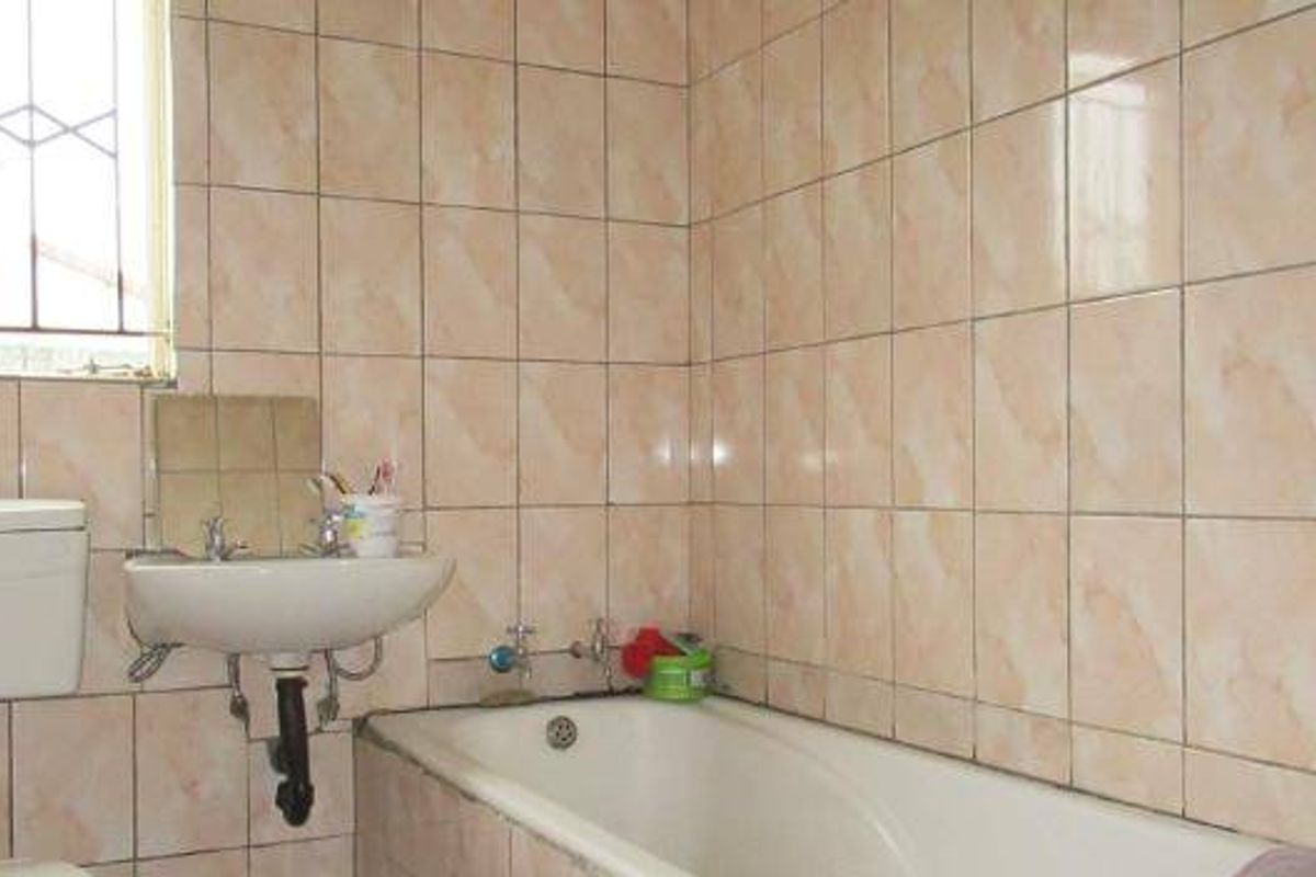 A dated bathroom featuring full-height wall tiling, a standard bathtub, wall-mounted basin, and a toilet cistern with security bars on the window.