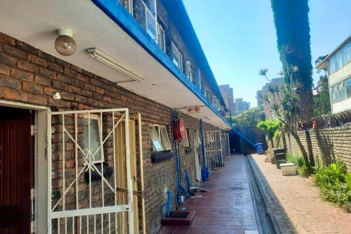 Exterior rear view of an apartment building featuring a tiled walkway, brick facade, and security infrastructure along the perimeter wall.