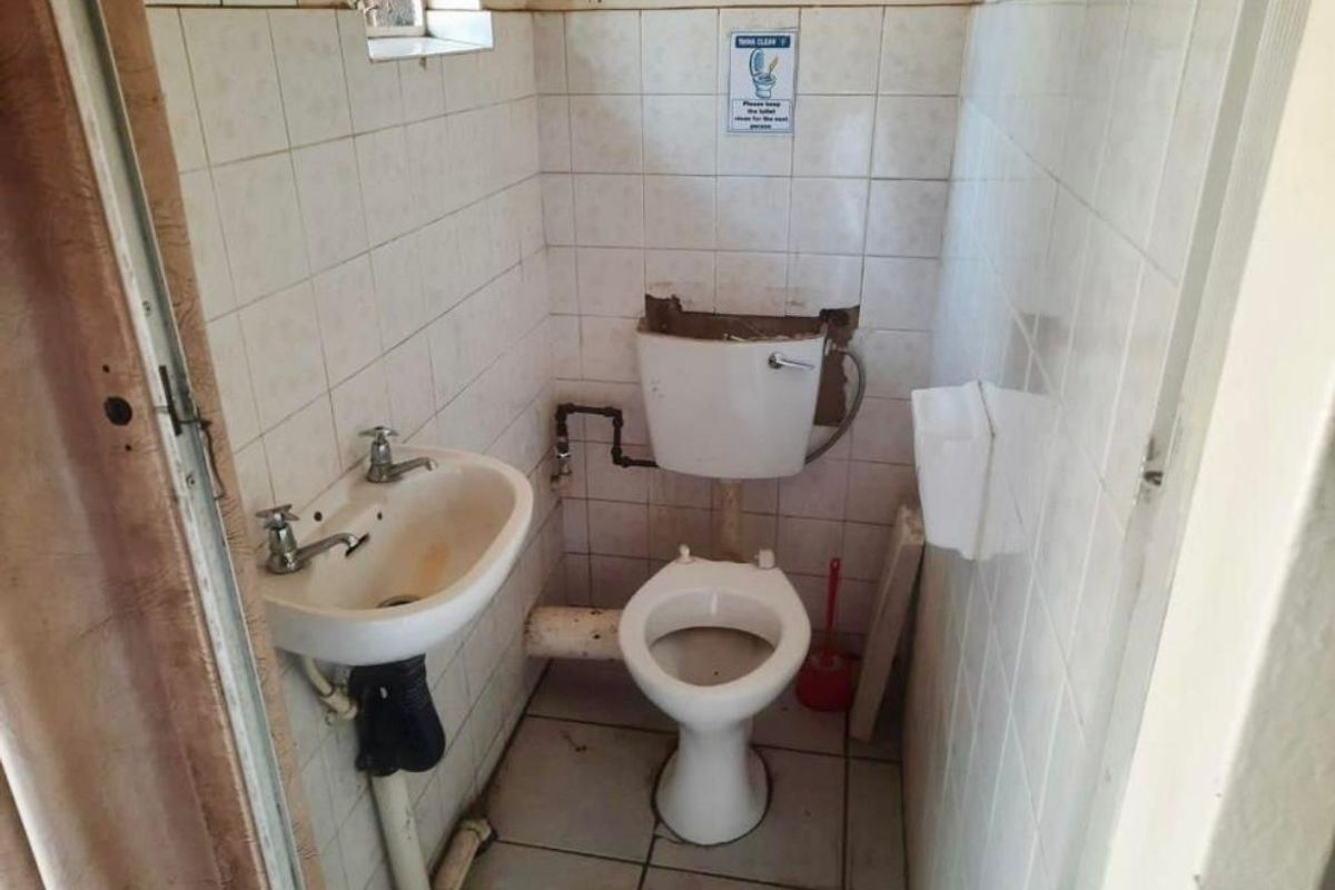 A small, dated bathroom featuring a pedestal basin and toilet with exposed plumbing and wall tiling in need of renovation.