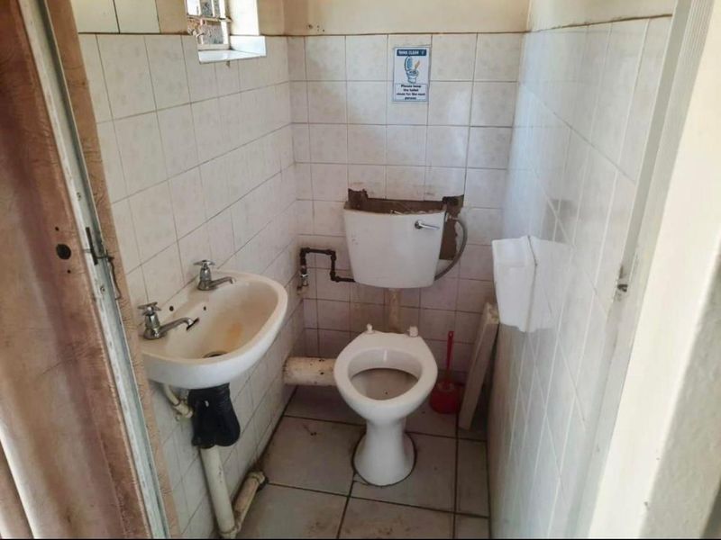 A small, dated bathroom featuring a pedestal basin and toilet with exposed plumbing and wall tiling in need of renovation.