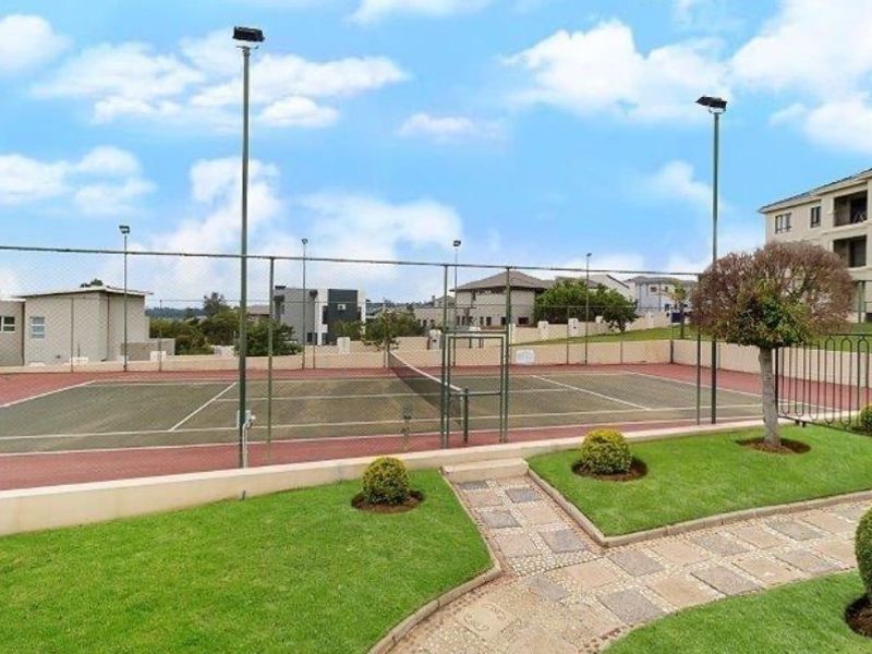 A well-maintained communal tennis court featuring floodlights, perimeter fencing, and surrounding landscaped garden areas within the apartment complex.