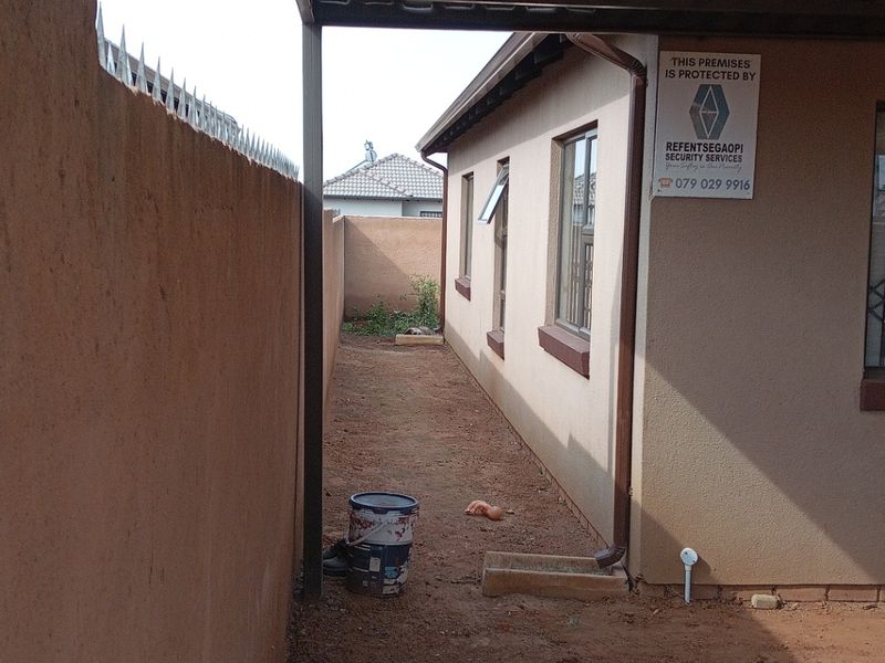 Side exterior area featuring a metal carport structure, perimeter wall with security spikes, and unpaved ground surface along the side of the house.