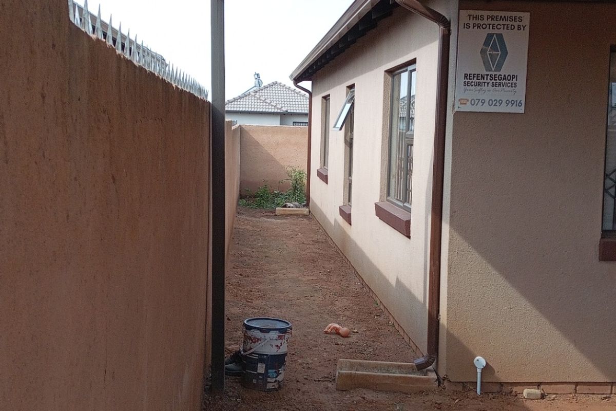 Side exterior area featuring a metal carport structure, perimeter wall with security spikes, and unpaved ground surface along the side of the house.