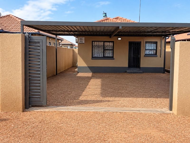 A well-maintained front exterior featuring a covered carport, secure gated entrance, and gravel driveway, enclosed by a solid perimeter wall.