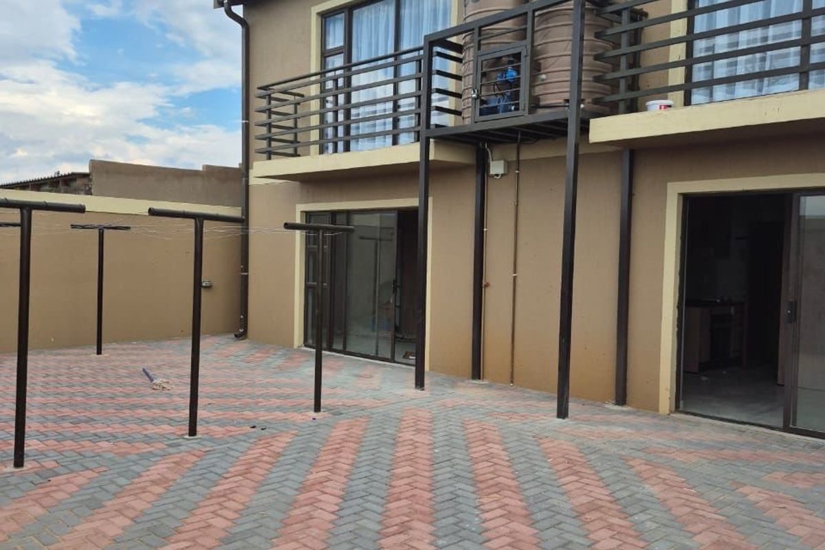 Rear exterior view of the apartment featuring a paved courtyard, a balcony with metal railings, and an integrated water storage tank system.
