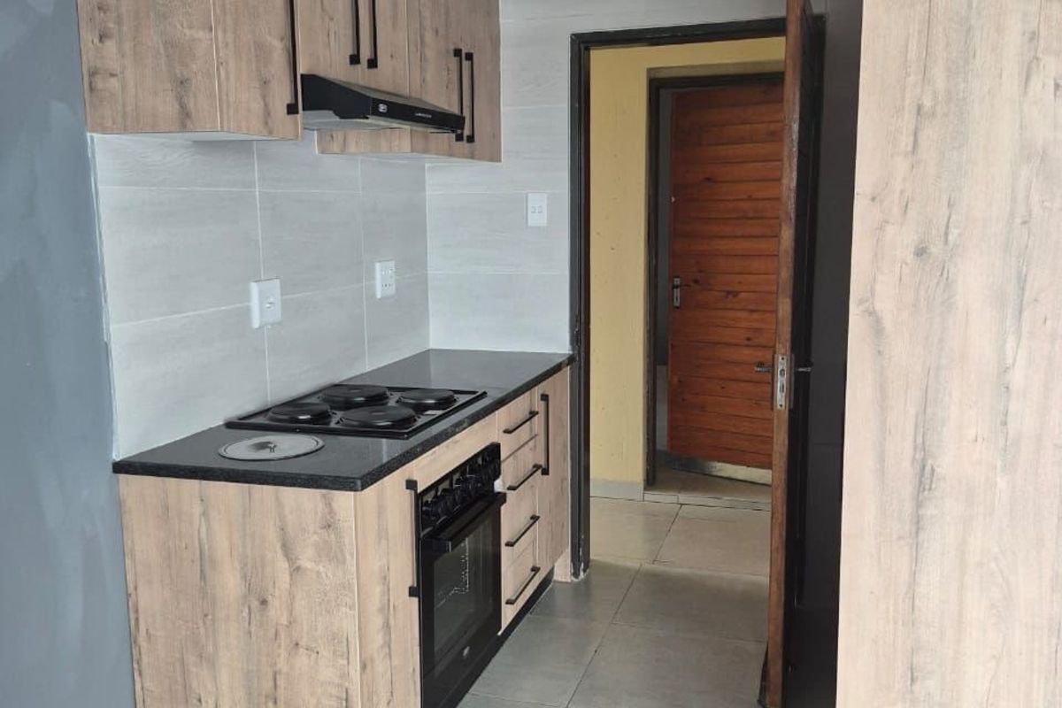 A modern, compact kitchen featuring wood-look cabinetry, dark countertops, integrated oven and hob, and neutral tiled flooring.
