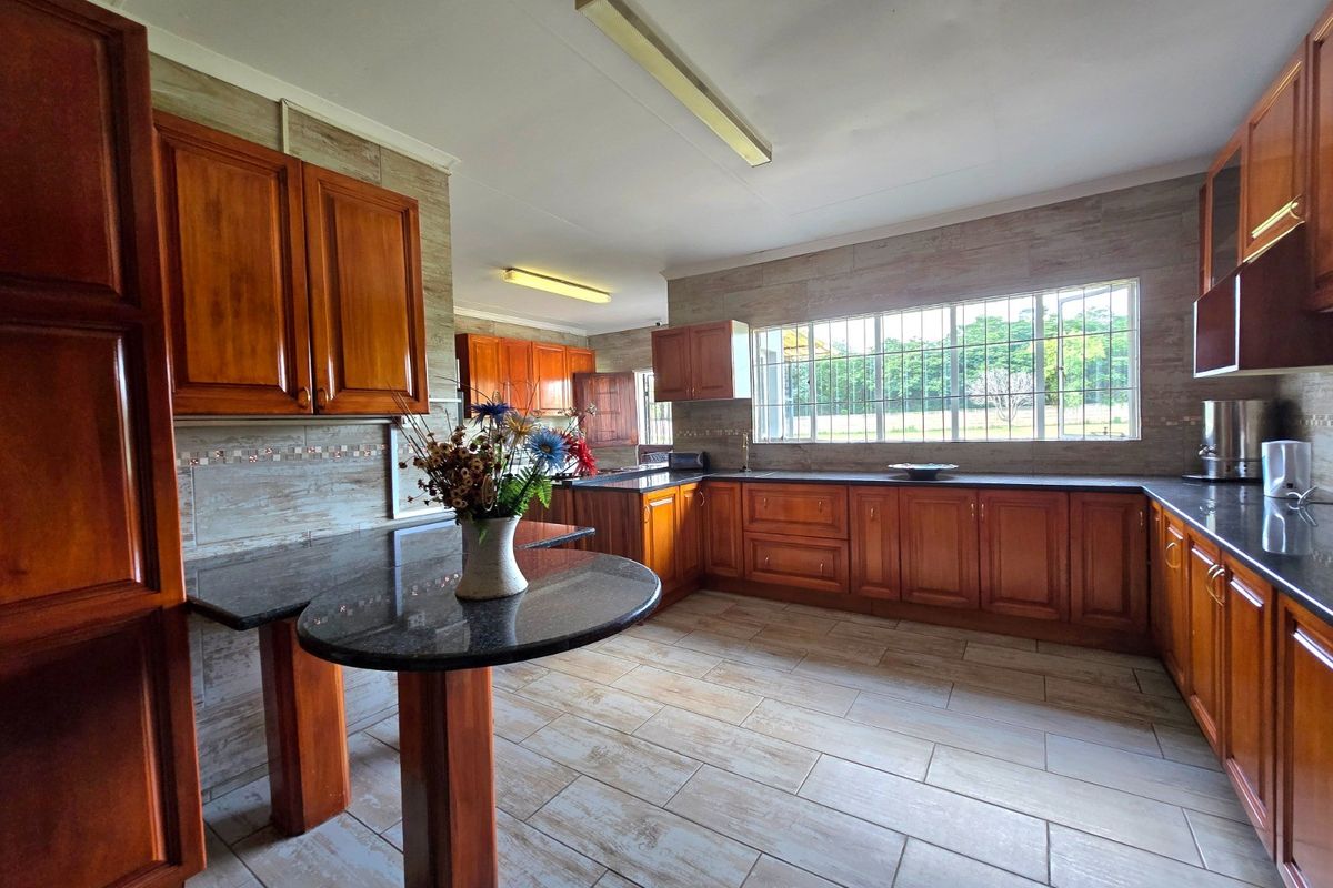Spacious kitchen featuring warm-toned wooden cabinetry, dark granite countertops, tiled flooring, and a large window with security bars overlooking the farm landscape.