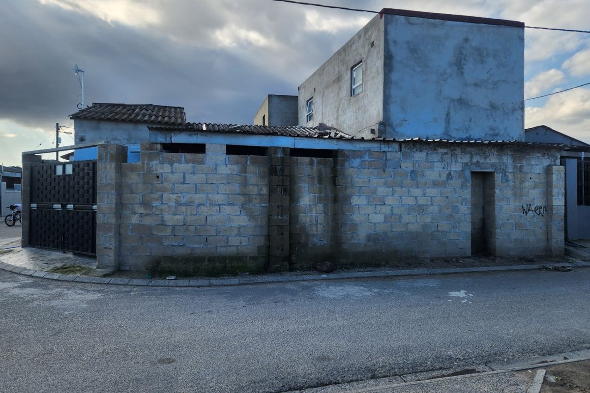 Street-facing view of a property featuring a concrete block boundary wall, metal gate, and multi-level structure with unfinished exterior plaster finishes.