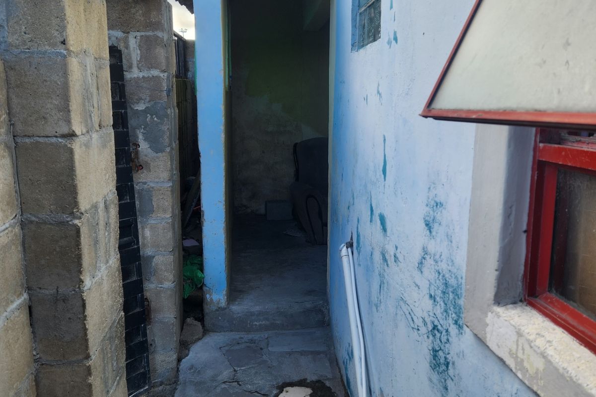 A narrow exterior side passage featuring a concrete block wall, weathered blue painted facade, and uneven paved flooring with exposed utility piping.