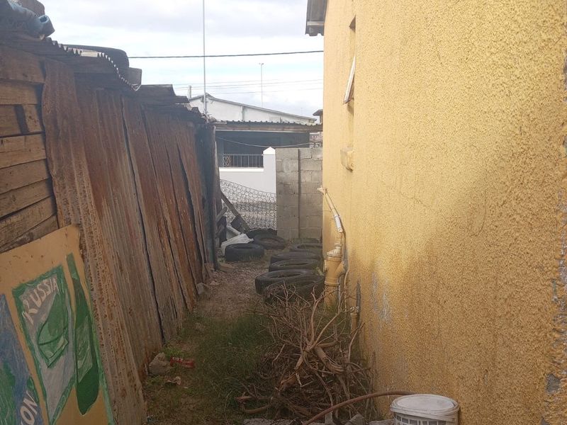 A narrow side passage between a plastered exterior wall and a corrugated metal fence, showing signs of wear and maintenance requirements.