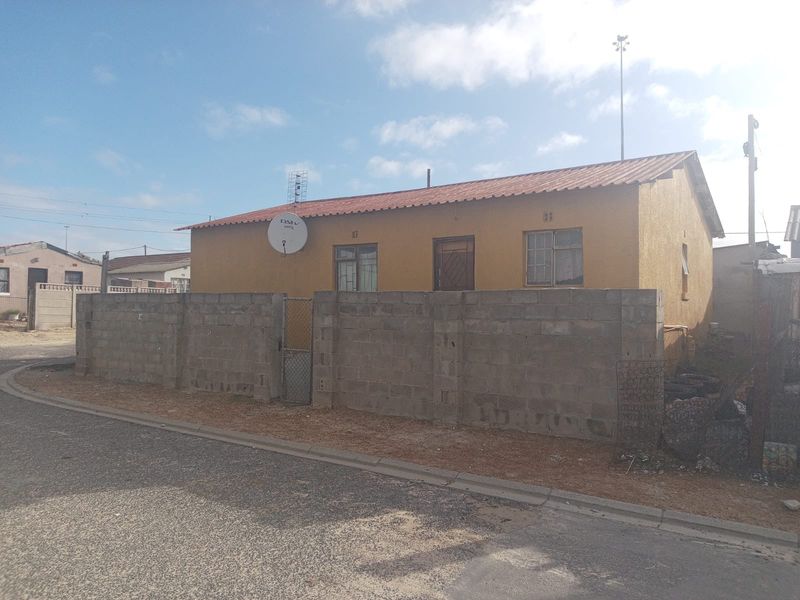 Front exterior view of a single-story house featuring a corrugated metal roof and a concrete block boundary wall with a metal gate.