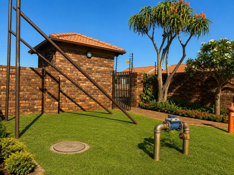 Front exterior view of the townhouse complex featuring a face brick boundary wall, electric fencing, security gate, and well-maintained landscaped garden area.