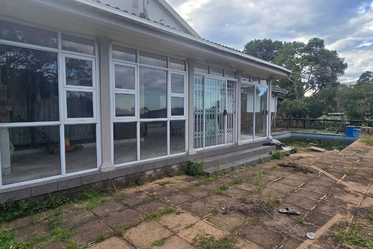 Rear exterior view featuring a paved patio area, large glass windows, sliding doors with security gates, and an outdoor swimming pool.