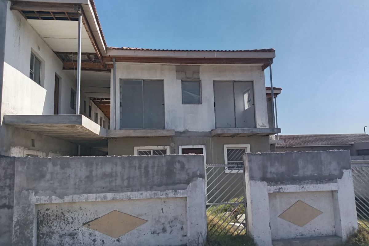 Front exterior view of a multi-story apartment building showing unfinished plaster, exposed roof eaves, and a concrete boundary wall with metal gate.
