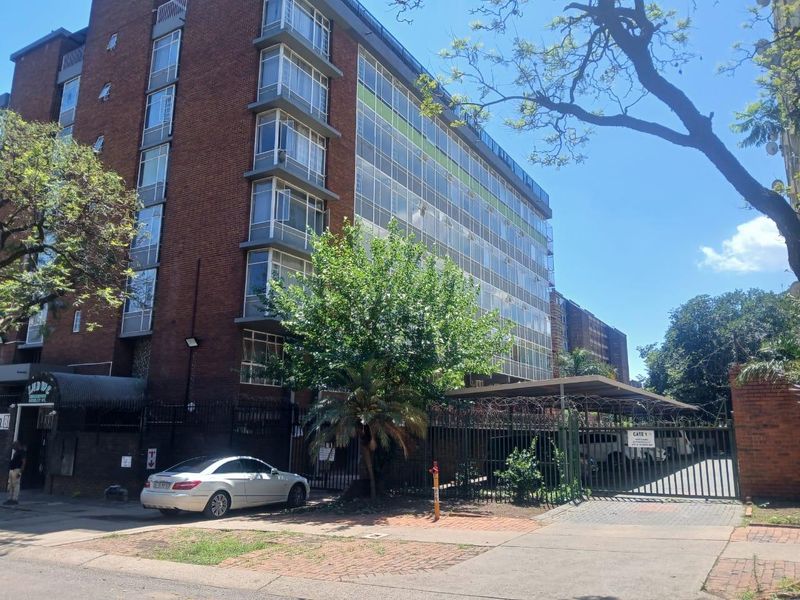 Street-level view of a multi-story brick apartment building featuring a gated entrance, carport parking, and security fencing.