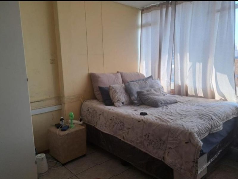 A bedroom featuring tiled flooring and a large window, currently showing signs of wear including wall staining and peeling paint.