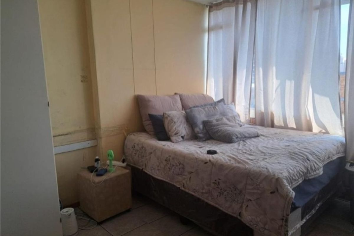 A bedroom featuring tiled flooring and a large window, currently showing signs of wear including wall staining and peeling paint.