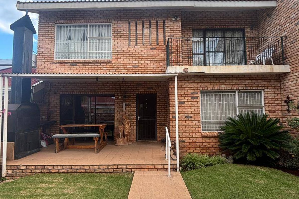 Rear view of a double-story brick townhouse featuring a covered patio with a built-in braai, a balcony, and a paved walkway through the garden.