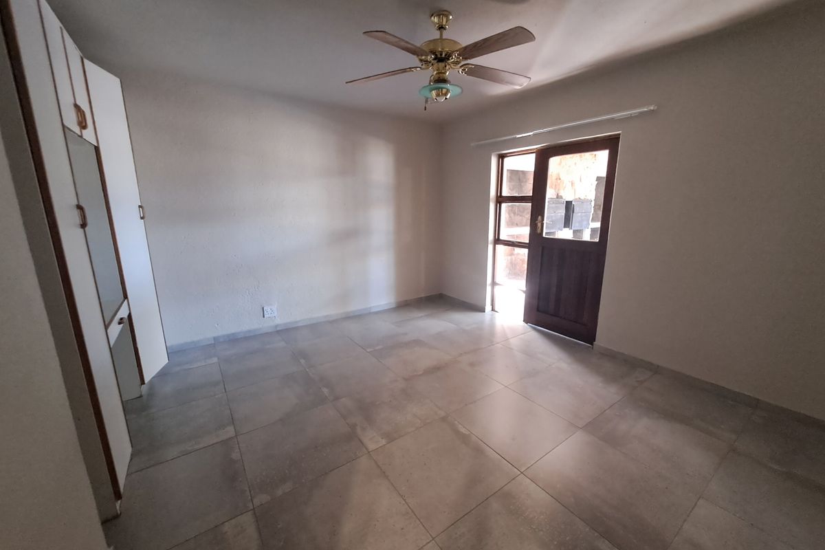 A spacious, empty bedroom featuring neutral tiled flooring, built-in cupboards, a ceiling fan, and direct access to an outdoor area.