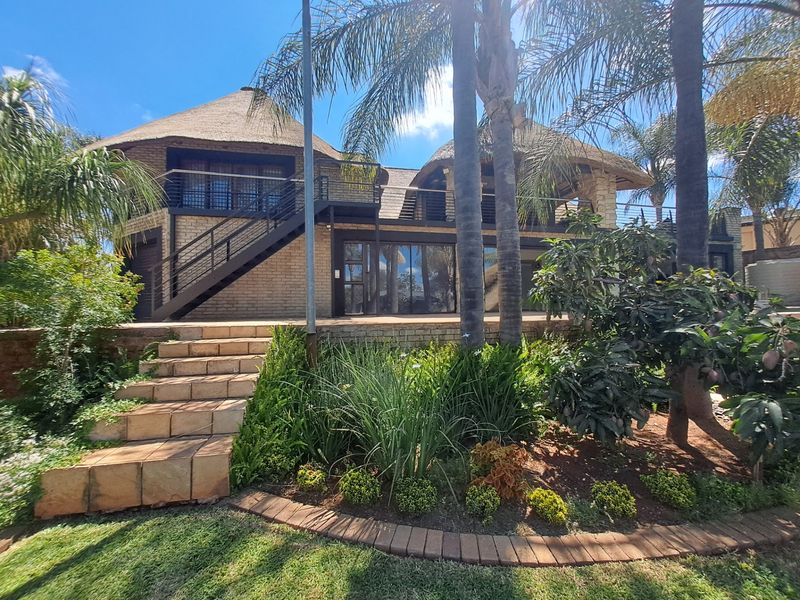 Rear view of a multi-level house featuring a thatched roof, brick exterior, metal staircase, and a balcony overlooking a landscaped garden with stone steps.