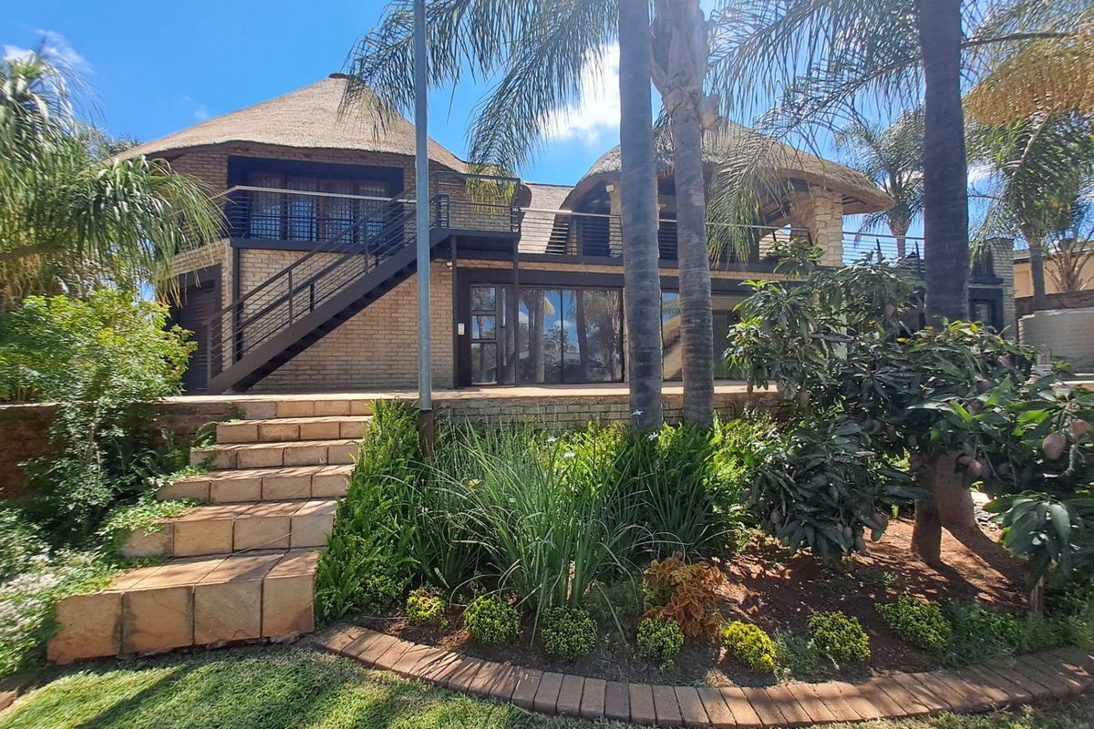 Rear view of a multi-level house featuring a thatched roof, brick exterior, metal staircase, and a balcony overlooking a landscaped garden with stone steps.