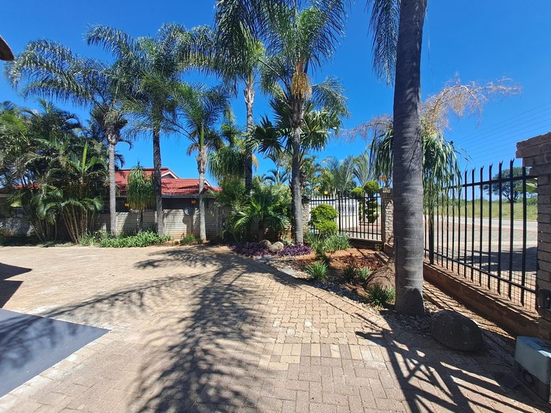 A well-maintained front exterior featuring a paved driveway, established palm trees, and secure metal perimeter fencing.