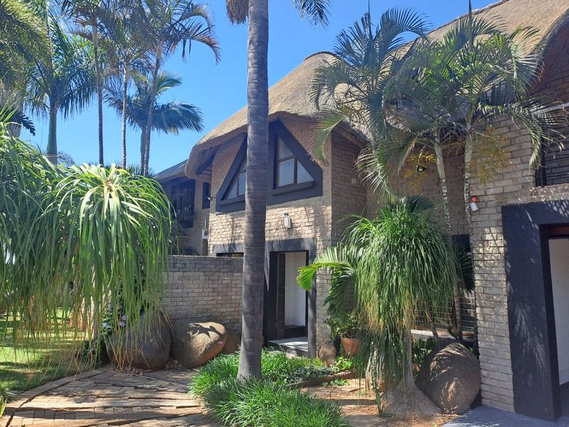 A charming face brick house featuring a traditional thatched roof, triangular architectural windows, and a well-maintained landscaped garden with a paved entrance path.