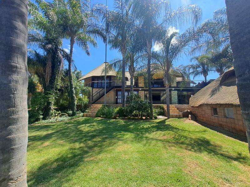 Rear view of a multi-level house featuring a thatched roof, stone facade, and a spacious landscaped garden with mature palm trees.
