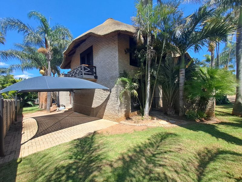 A two-story brick house featuring a traditional thatch roof, a paved driveway with a shade sail, and established garden landscaping.