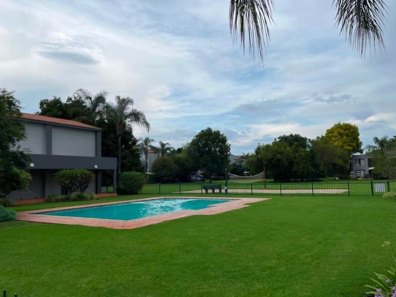 A well-maintained communal pool area featuring a swimming pool with brick coping, surrounded by expansive green lawns and a fenced volleyball court.