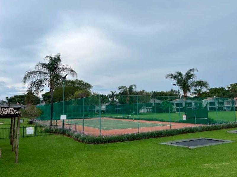 Well-maintained communal tennis court with perimeter fencing and floodlights, situated within a landscaped residential estate area featuring an in-ground trampoline.