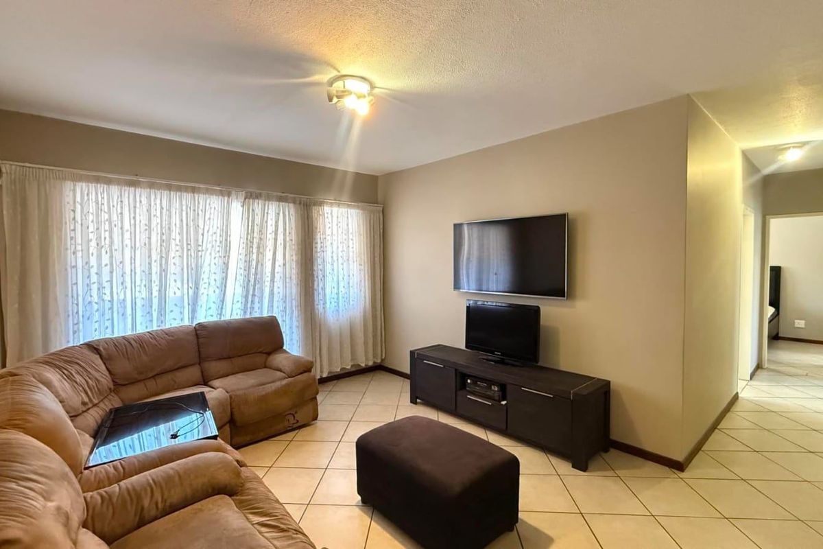 A comfortable living room featuring neutral wall tones, tiled flooring, and large windows with curtain treatments, leading into a hallway.