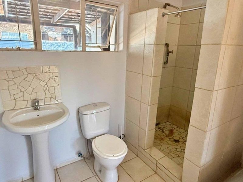 A dated bathroom featuring a pedestal basin, toilet, and a tiled shower enclosure with visible wear and tear on the floor and fixtures.