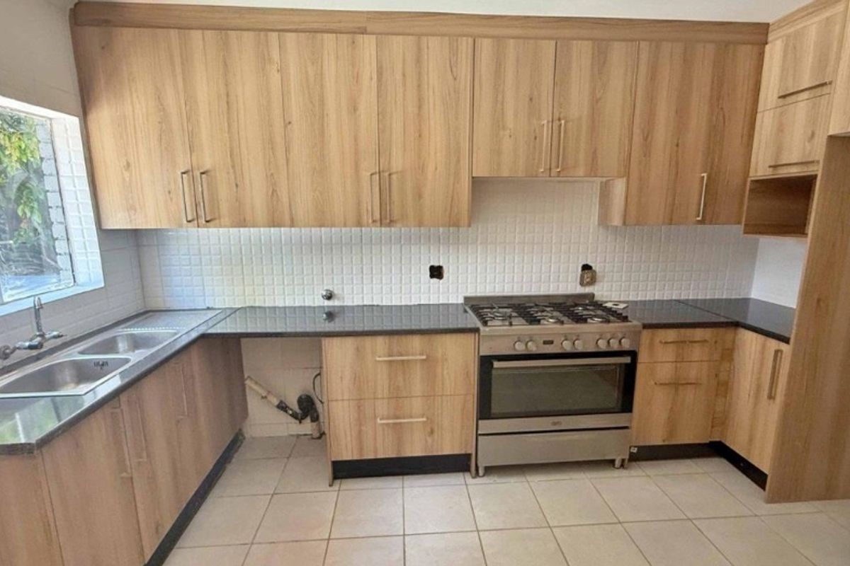 A functional kitchen featuring wood-look cabinetry, dark granite countertops, a stainless steel gas stove, and a double sink with tiled backsplash.