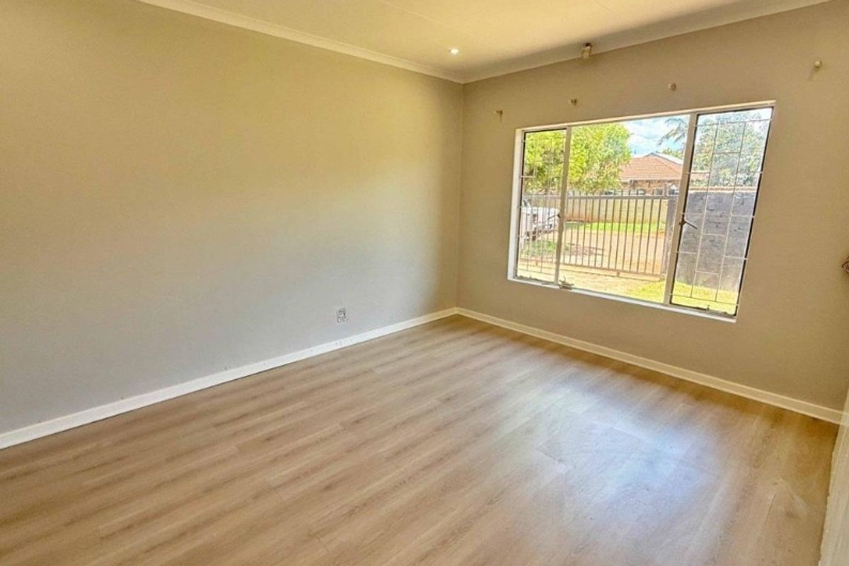 A bright, empty bedroom featuring light-toned laminate flooring, neutral walls, and a large window with security bars overlooking the front yard.
