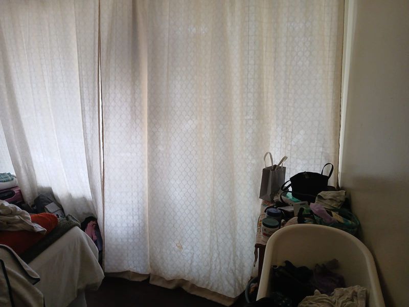 A bedroom interior featuring floor-to-ceiling curtains, currently cluttered with personal items and showing signs of wear.