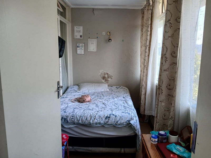 A compact bedroom featuring large windows for natural light, currently showing signs of wall surface damage and plaster cracking requiring maintenance.