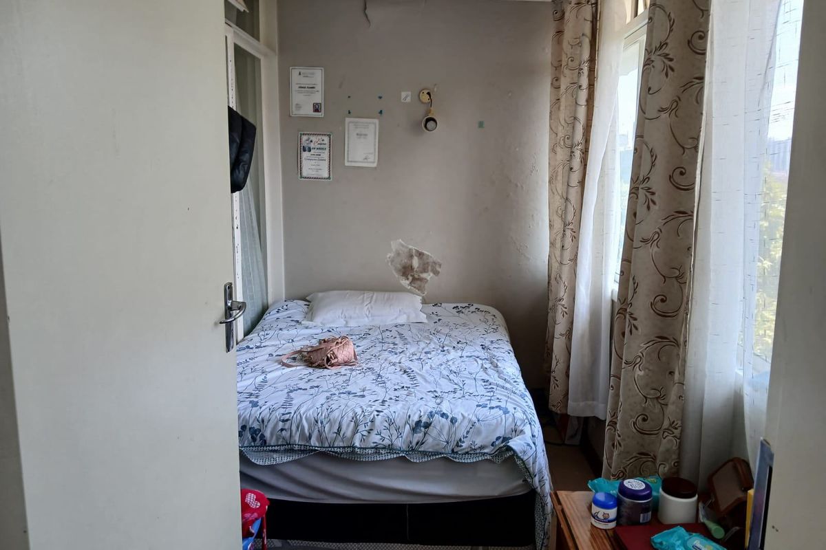A compact bedroom featuring large windows for natural light, currently showing signs of wall surface damage and plaster cracking requiring maintenance.