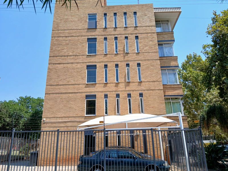 A multi-story apartment building facade featuring face-brick construction, vertical casement windows, and secure perimeter fencing with an integrated carport.