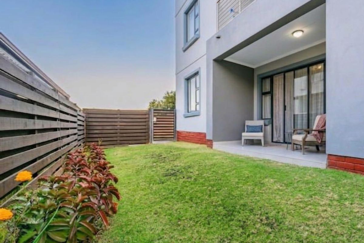 A private rear garden featuring a well-maintained lawn, timber fencing, and a covered patio area with sliding door access to the apartment interior.