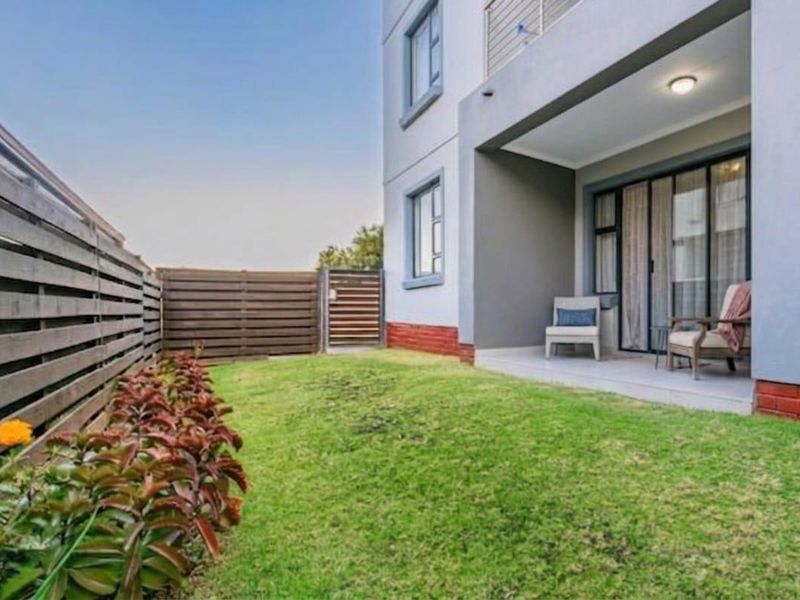 A private rear garden featuring a well-maintained lawn, timber fencing, and a covered patio area with sliding door access to the apartment interior.